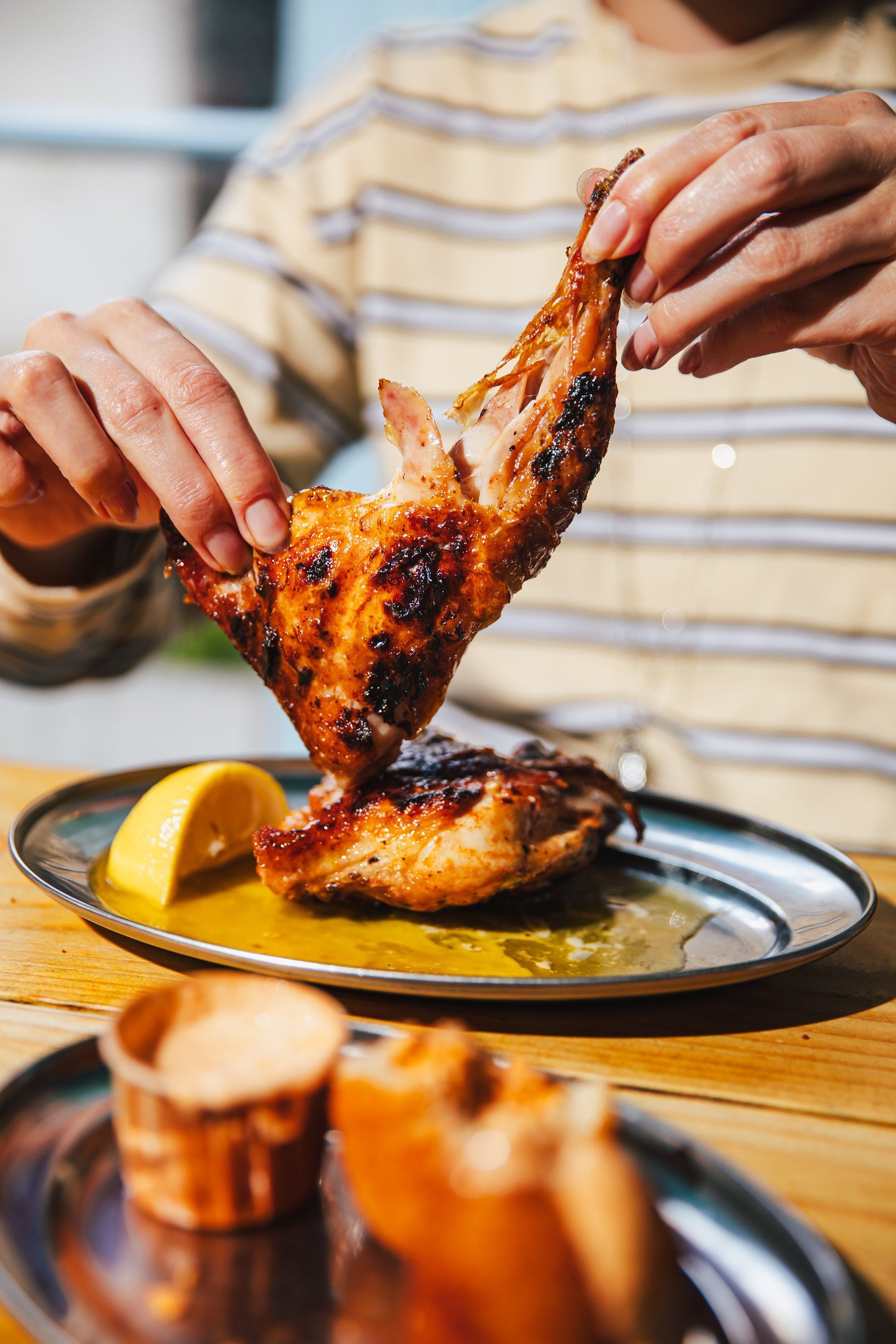 Hands tearing a flame-grilled half chicken over a plate with lemon and piri-piri sauce
