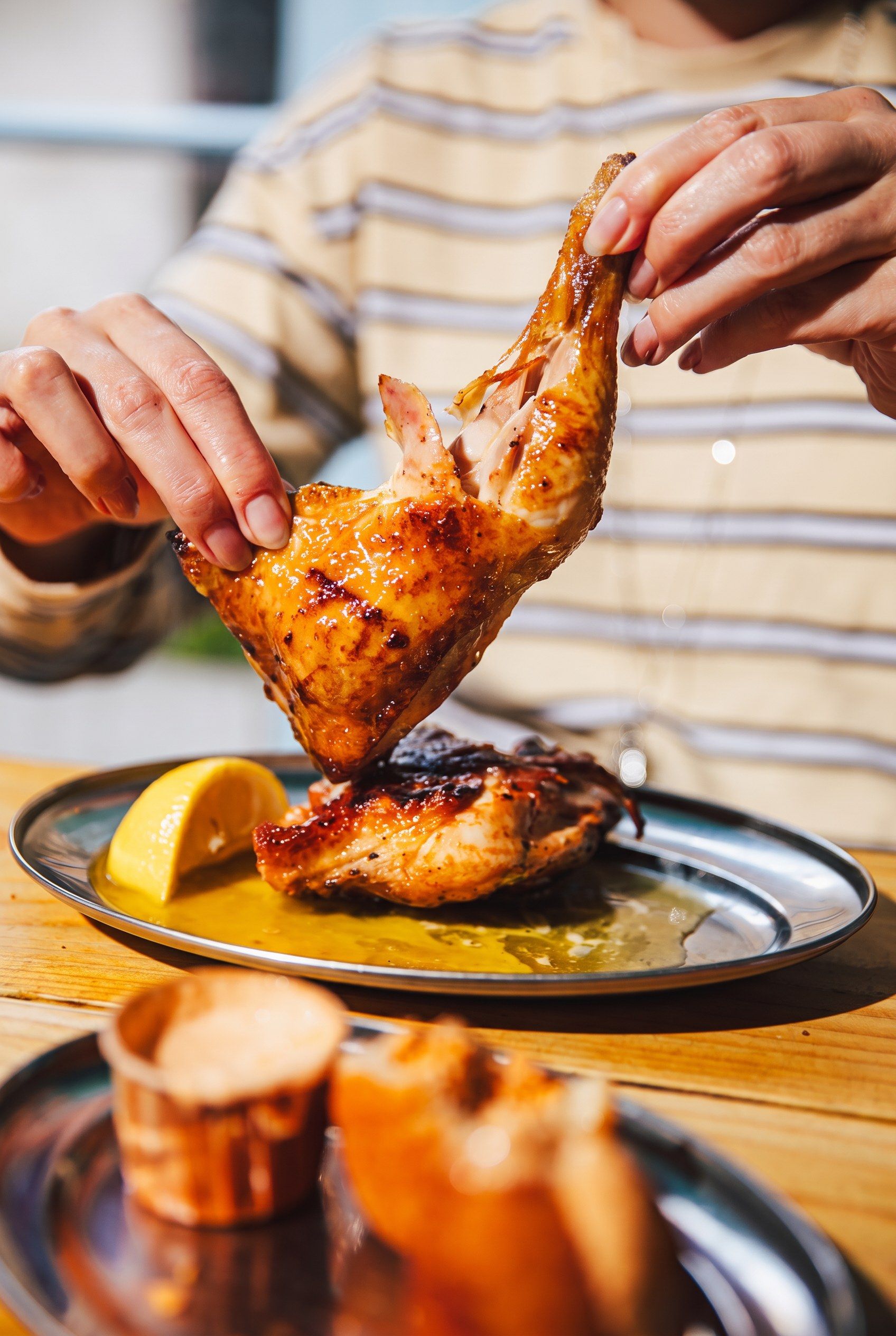 Hands tearing a flame-grilled half chicken over a plate with lemon and piri-piri sauce